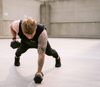 A fit man wearing a black Aion weighted vest performing a single-arm dumbbell row in a plank position outdoors, showcasing core stability, strength training, and functional fitness performance.