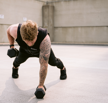 A fit man wearing a black Aion weighted vest performing a single-arm dumbbell row in a plank position outdoors, showcasing core stability, strength training, and functional fitness performance.