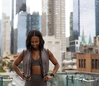 fit young woman smiling and standing on a balcony with New York City behind her wearing the Aion Women's Weighted Walking Vest